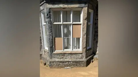 Christopher Beaumont A window in a stone wall has eight panes of glass. Two of them have been boarded up neatly.