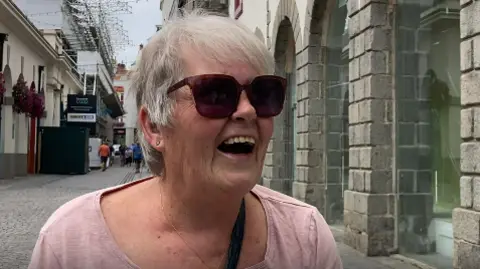Close-up of Trish Smith with an open-mouthed smile. She has short, light grey hair and is wearing sunglasses and a pink top. There is a street scene behind her.