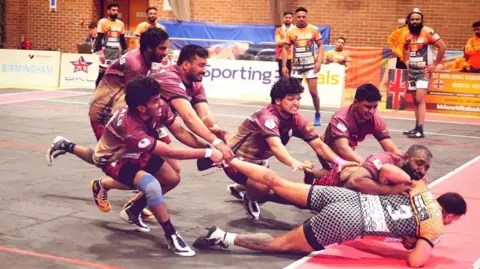 British Kabaddi League A Wolverhampton raider is halted in his tracks during a British Kabaddi League match. Six members of the opposing team are grabbing his arm and leg to stop him crossing the line.