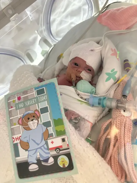 Caitlin Duncan A little baby with a white hat on in a cot with tubes going up her nose and a card which says "I had surgery today" on it