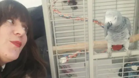 Contributed A woman with dark brown hair leans toward a parrot sitting in a white cage. She has long brown hair and wears red lipstick. She looks toward the parrot. The parrot is grey with some white and red feathers. 