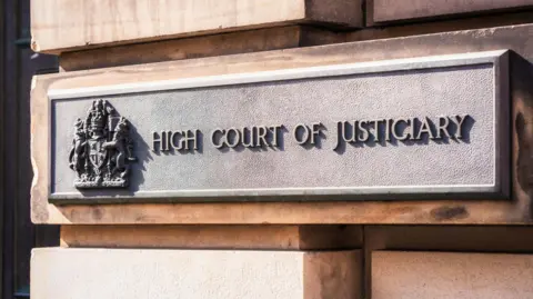A metal sign on a sandstone block exterior wall. The sign has a royal crest and the words "high court of justiciary".