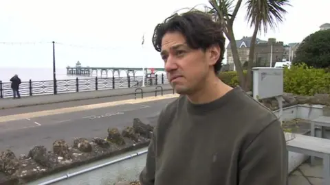 A man looking sad stands in the garden of his restaurant which overlooks the seafront.