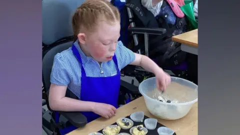 Ridgeway School Amelia sits at a desk in school wearing her checked blue dress and a royal blue apron. She is making cakes and has her hand in a clear plastic bowl, picking out blueberries. Her auburn hair is tied back in a French plait and her cheek is flushed.