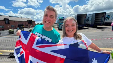 Tony Hamlyn and Lynsey Richmond posing with an Australia flag and smiling big grins. It is sunny and there are four trucks behind them. Hamlyn is wearing a green top and has gelled up hair. Richmond has a white T-shirt and glitter on her face. 