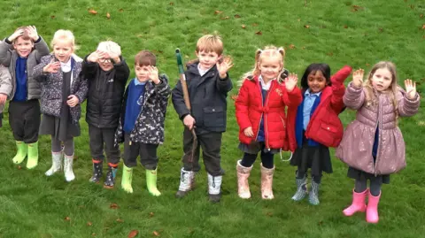 An aerial view of a group of primary school children on a green field waving at the camera  during a tree-planting event. They are wearing wellies and colourful coats and one holds a small garden fork.