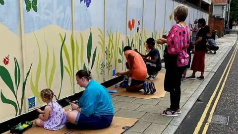 Stephanie Northen Children and adults paint a mural on a hoarding outside a building, drawing plants and butterflies upon a blue background