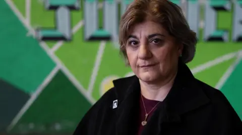 Reuters Kimia Zabihyan, a woman with short brown hair wearing a black fleece and maroon top, stands in front of a green mural that says 'justice'.