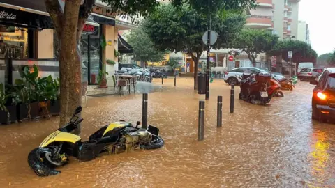 EPA Una calle inundada de agua fangosa en la ciudad de Ibiza cuenta con una moto amarilla que cae de lado y algunos autos que conducen a través del agua marrón.