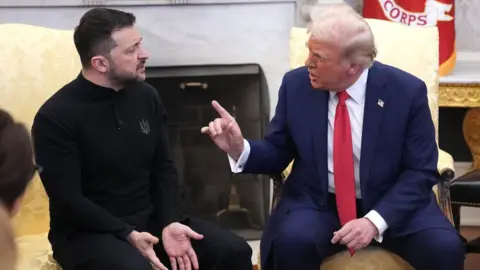 Getty Images President Zelensky, with a dark beard and hair and wearing all black, sits next to Donald Trump, with greying hair and wearing a blue suit and red tie. Trump, on the right, has his finger pointed at Zelensky, who has his arms pointed down with his palms turned over towards the camera. 