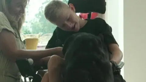 A blonde boy in a wheelchair smiles and leans down towards a large black dog.