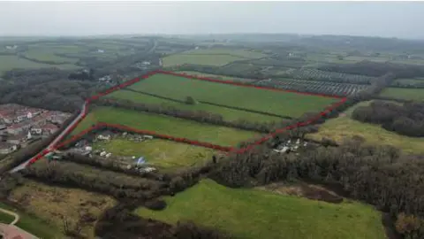 An aerial photo of a large patch of land that is square in shape with a thin section stretching off from one corner. The land is agricultural land with a large hedge running through the centre.