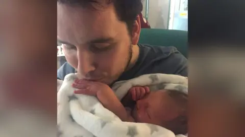 Michael with black hair kissing the hand of his newborn baby Anna, wrapped in a white blanket with grey stars.