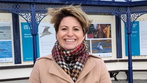 Anttoni James Numminen A woman with short blonde hair smiles at the camera. She wears a camel- coloured coat and a black, red and brown checked scarf. 