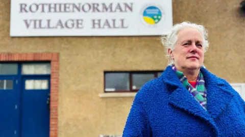 Elaine Bryson standing in front of Roithienorman Village Hall sign, she has grey hair and is wearing a fleecy dark blue jacket.