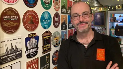 Graham Newbury standing next to a wall inside. The words Bridge Street Ale House are in yellow on an orange background on his black top.