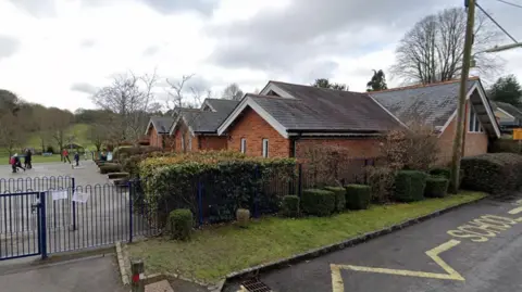 Single story pitched roof brick building with blue metal fence and hedging a playground with children running around is to the left of the building with fields seen beyond.