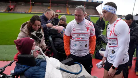 PA Media Dan Allinson, who has MND and is in a wheelchair, laughing with Sinfield alongside the pitch at Craven Park stadium, surrounded by other members of the MND community and supporters 