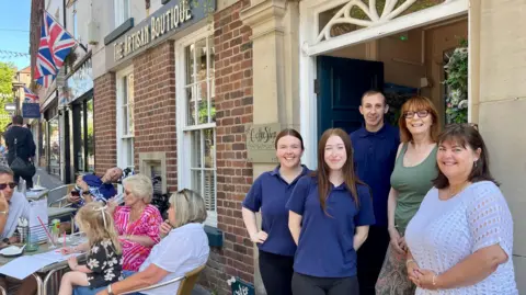 Four women and one man stand in the doorway of a cafe. Outside, people are sat at outdoor tables. The sign above the face reads The Artisan Boutique.