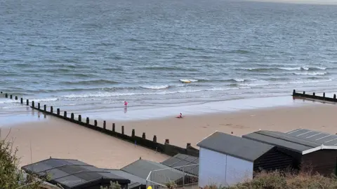 Simon Dedman/BBC The beach in Frinton, which has wooden sea defences and beach huts. Two childen play on the sand in a reasonably high tide.