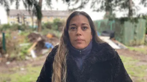 Kelly Harryman, a woman with long brown hair wearing a black coat, stands in front of a pile of rubbish dumped in a cemetery. 