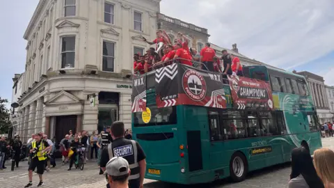 The players are on the bus looking out of the back of it at the crowd. There are security officers standing near the bus.