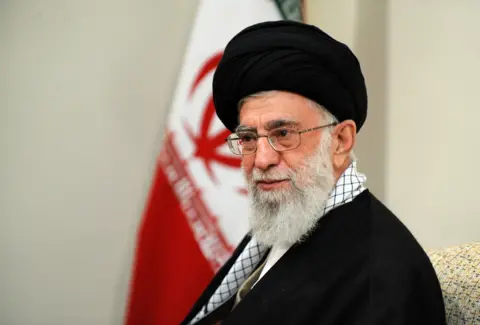 Getty Images Ayatollah Ali Khamenei, who has a long grey bears an glasses, sits side on in front of a wall and a large flag. He is wearing a black robe and head piece. 