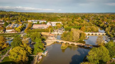An aerial view of the centre of Stratford-on-Avon with trees on both sides of a wide river and large buildings seen between them. There are two large bridges crossing the river 
