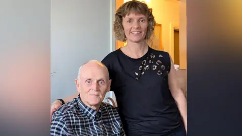 An elderly man in a buttoned up shirt sits next to a younger woman with fair curly hair who stands at his side.