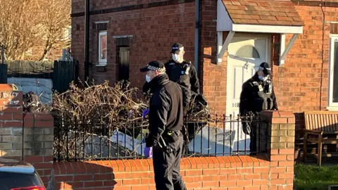 Three police officers are standing in a garden and on the path. Thy are dressed in black, wearing masks and police caps.