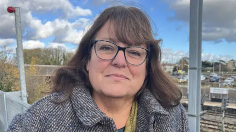 Nicola Haseler/BBC A woman looking at the camera, wearing glasses and a woollen coat. There are railway lines and a platform behind her.