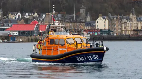 Bright orange Oban lifeboat pictured moving through Oban Bay with the town of Oban in the background.