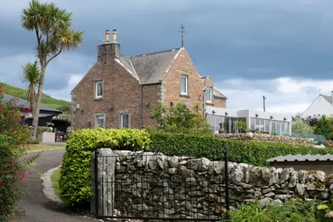 IWC Media A traditional stone house with a pitched roof sits behind a low stone wall and iron gate, surrounded by lush green shrubs and garden plants.
A tall, spiky tree stands to the left, while a modern extension with large windows is visible on the right side of the building.
Dark, heavy clouds fill the sky above, contrasting with the warm tones of the stone exterior and the greenery below.