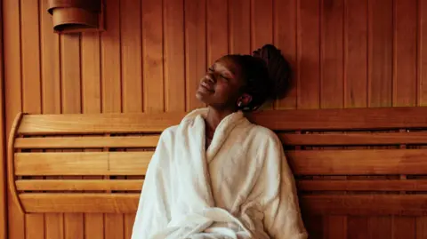 A woman wearing a white dressing gown sits in a room with wooden walls. She has her eyes closed and is smiling.