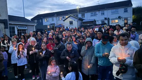 A crowd of about 100 people all wearing coats of different colours and many holding candles stand near the victim's home.