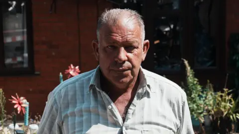 John Fairhall/BBC Mark Matthews a man standing in front of a property in Kenninghall. The property is cordoned off following a significant fire which has left the property inhabitable. Matthews is looking directly at the camera and is wearing a life patterned shirt.