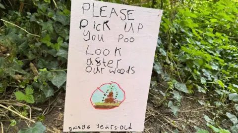 Family handout A sign sticking in the ground in the woods telling people to pick up their poo. 
