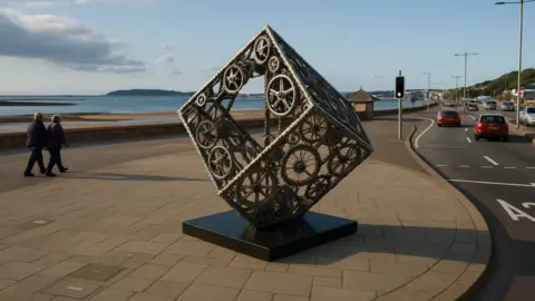 A picture of the sculpture on the seafront. It is a square shaped design leaning at an angle.