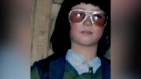 Zahra Joya A close-up, slightly blurry shot of Zahra dressed as a boy. She has thick black chin-length hair with a heavy fringe, and wears aviator style sunglasses. She has a white shirt and a blue waistcoat.