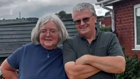 A woman with bobbed grey hair and a man with short grey hair are smiling while standing next to each other. They are on decking, with railing and a house behind them.