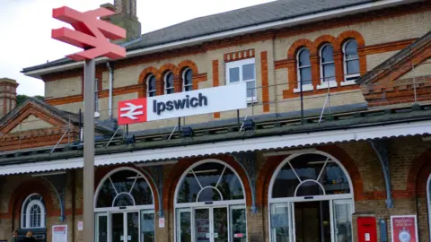Greater Anglia The entrance to Ipswich train station. The light and dark brick building has rounded windows and a large red and white Ipswich sign on the front.