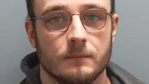 In a police custody image, Bradley Talbot is pictured with glasses, receding brown hair and a short beard.