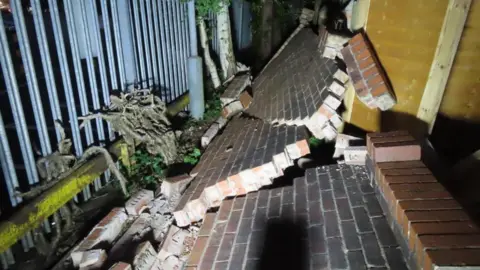 A collapsed wall with large lumps of brick and cement overs the walkway. On the left is a metal security fence and there is wooden fence panelling on the right of the picture.