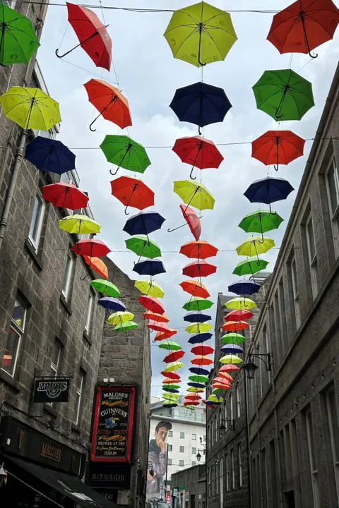Brian Carroll Multi coloured umbrellas hang from an art canopy over a street in Aberdeen. There is a mural of a man at the far end on another building.