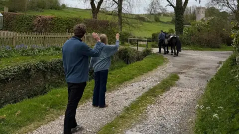 Roger Sewill Two people waving to Roger Sewill and his pony Scarlet during their trek. It is a cloudy day.