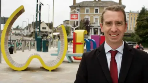 Peter Taylor with short brown hair, wearing a black jacket over a white shirt and red tie. There is play equipment in the background painted in bright colours. There is a railway station sign behind, and a three-storey wood-faced building beyond that.