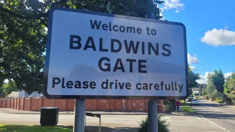 A white road sign with black letters that read 'Welcome to Baldwins Gate please drive carefully' is seen positioned at the side of a road junction.