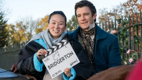 A man and woman together outdoors holding a film clapperboard that reads ‘Bridgerton’ along with scene and take numbers. The man is wearing period dress. There is a green metal fence or railings behind. It is sunny.