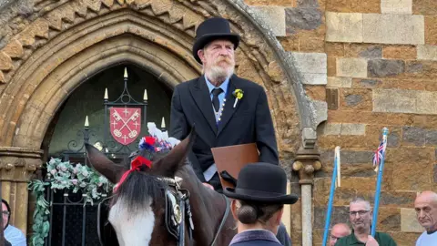 Carroll Weston/BBC The bailiff is on horseback holding a brown folder. He is wearing a black jacket and a black top hat. 