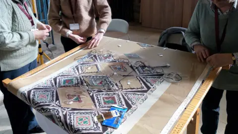 Peterborough Cathedral A white cloth with colourful patterns of stitching on it. Three people can be seen standing around it.